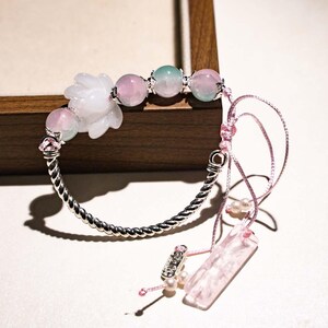 Puede incluir: Una pulsera delicada con una banda de metal retorcido plateado. Cuenta con cuentas de piedras preciosas rosas y verdes, un dije en forma de flor blanca y una borla rosa con un dije rectangular. La pulsera está sobre una superficie blanca.