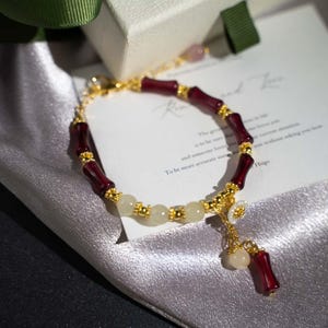 May include: A gold-toned bracelet featuring alternating burgundy and cream-colored beads. The bracelet has a delicate design with a dangling charm and is set against a satin background. The bracelet is a piece of jewelry.