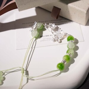 May include: A delicate bracelet featuring green and white gemstone beads. The bracelet is accented with silver-colored floral and butterfly charms. The beads are strung on a light green cord, and the bracelet is displayed on a white surface.