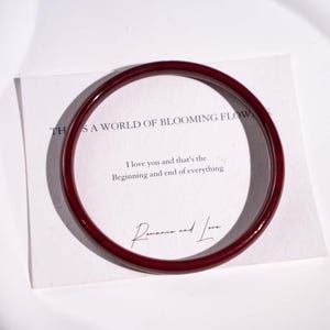 May include: A deep red bangle with a shiny surface lies on a white card. The card displays the text "THIS IS A WORLD OF BLOOMING FLOWERS" and a romantic quote. The words "Romance and Love" are also visible.
