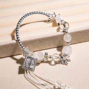 Puede incluir: Una pulsera plateada con cuentas blancas y abalorios decorativos. La pulsera presenta una banda de metal retorcido y un dije cuadrado con caracteres chinos. Delicadas borlas plateadas cuelgan del dije.
