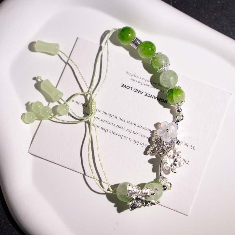 May include: A green beaded bracelet with silver accents and floral charms. The bracelet features a mix of light and dark green beads, with a delicate string closure. The text on the card reads "ROMANCE AND LOVE".