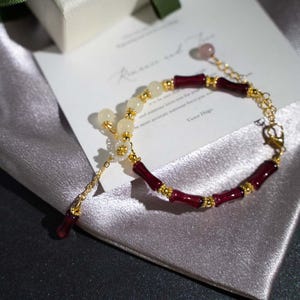 May include: A gold-toned bracelet with alternating sections of burgundy bamboo-shaped beads and creamy white beads. The bracelet includes a delicate chain and a small, dangling burgundy bead. A card with text is in the background.