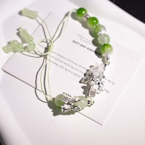 May include: A delicate green beaded bracelet with silver floral accents. The bracelet features round, light green beads and silver flower charms. The text on the card reads "ROMANCE AND LOVE".