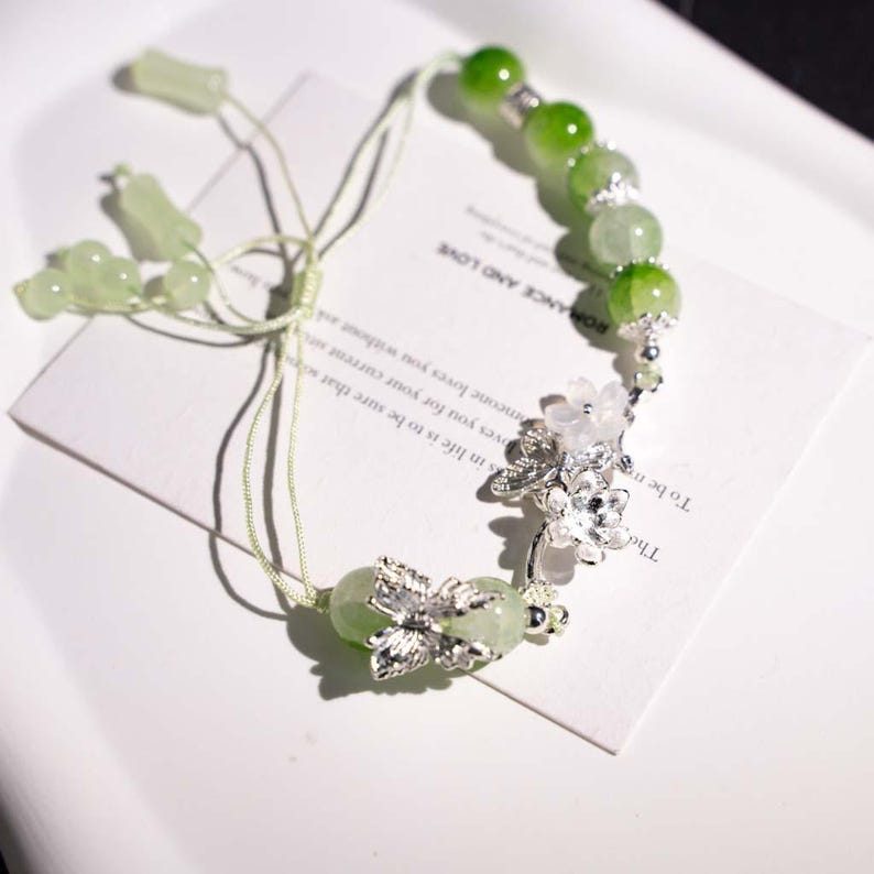 May include: A delicate green beaded bracelet with silver floral accents. The bracelet features round, light green beads and silver flower charms. The text on the card reads "ROMANCE AND LOVE".
