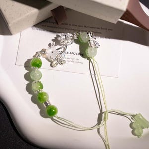 May include: A delicate bracelet featuring green and white beads, accented with silver-toned floral and butterfly charms. The bracelet has a light green cord and a decorative charm at the end. The text "MACE AND LOVE" is visible on a card in the background.