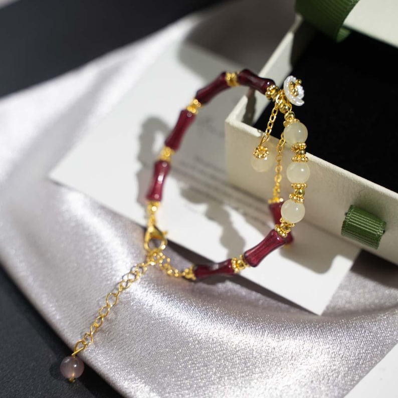 May include: A gold-toned bracelet with burgundy bamboo-shaped links and white spherical beads. The bracelet features a delicate chain with a small, round, purple charm. A small white flower charm hangs from the bracelet.