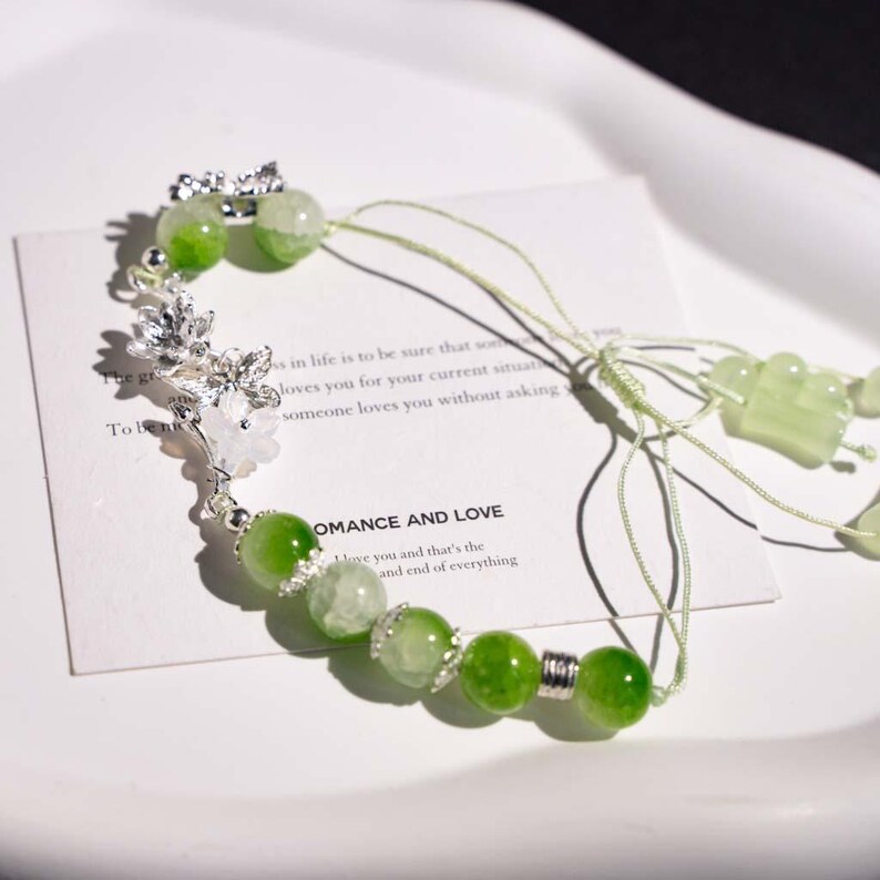 May include: A delicate bracelet featuring green and clear beads, accented with silver-toned floral charms. The bracelet includes a card with the text "ROMANCE AND LOVE" and other text. The bracelet is on a white surface.