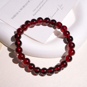 May include: A bracelet made of polished, deep red beads. The beads are round and strung together to form a circular shape. The bracelet is displayed on a white surface with a piece of paper that reads "THIS IS A WORLD OF" and "I love you and that's Beginning and end of everything."