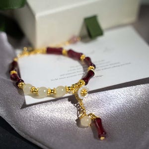May include: A gold-toned bracelet featuring alternating burgundy and cream-colored beads. The bracelet has a delicate floral charm and a dangling accent. The design incorporates small gold-colored accents throughout the bracelet.
