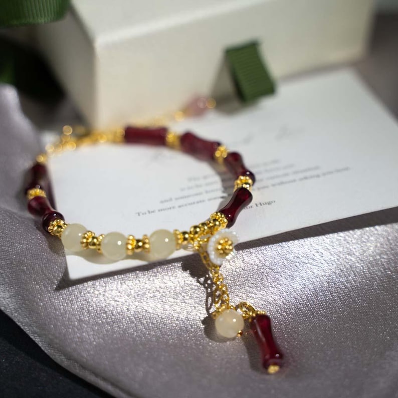 May include: A gold-toned bracelet featuring alternating burgundy and cream-colored beads. The bracelet has a delicate floral charm and a dangling accent. The design incorporates small gold-colored accents throughout the bracelet.