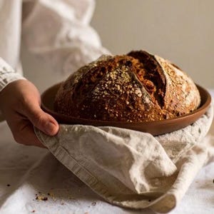 以下が含まれることがあります： 焼き立ての丸い形のパン。 表面は黄金色で、素朴な外観。 木製の皿にのせられ、リネンの布の上に置かれています。 この画像は、温かさと手作りの良さを感じさせます。