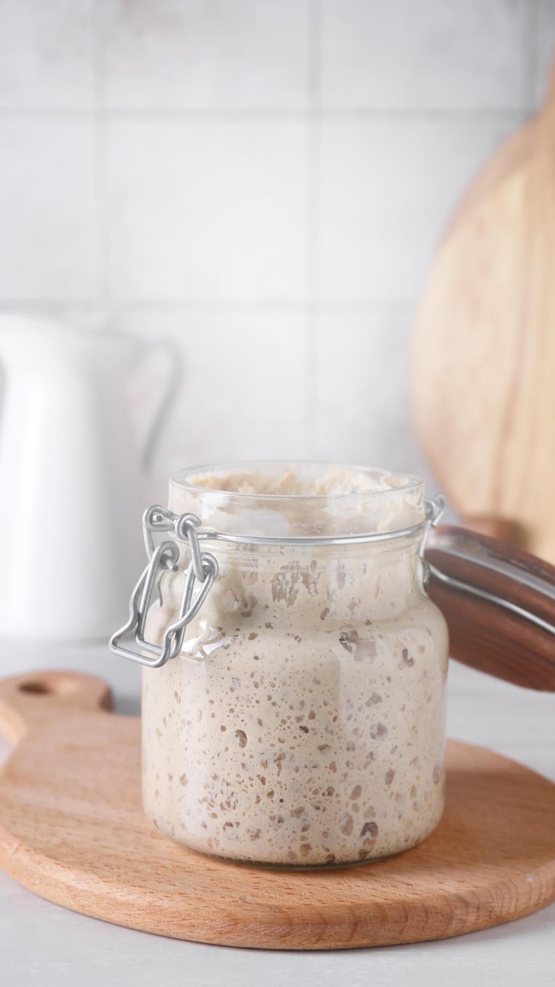 Könnte beinhalten: Ein klares Glas mit Metallverschluss, gefüllt mit einer blasenreichen, hellen Mischung, wahrscheinlich Sauerteigstarter. Das Glas steht auf einem Holzschneidebrett, mit einer weißen Kanne und einem Brett im Hintergrund.