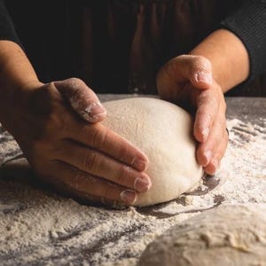 以下が含まれることがあります： 粉をまぶした表面で生地の塊をこねている手のクローズアップ。生地は薄いベージュ色で、手には小麦粉がまぶされています。別の生地の塊が前景に見えます。