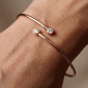 Op de afbeelding: Een open armband van roségoud met twee ronde, heldere, gefacetteerde stenen in een zetting aan elk uiteinde van de dunne, gepolijste metalen band. De armband is ontworpen met een opening tussen de twee stenen voor een strakke uitstraling.