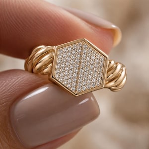 May include: A gold ring featuring a hexagonal face encrusted with small, sparkling stones. The ring's band has a twisted rope design on each side of the hexagonal face. The ring is held between two fingers.