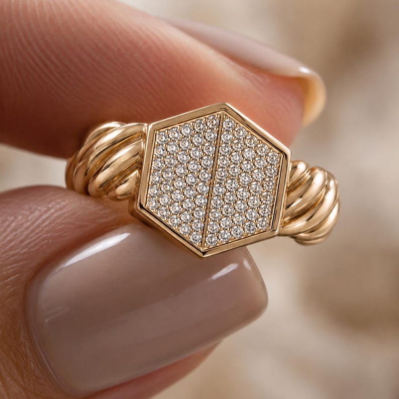 May include: A gold ring featuring a hexagonal face encrusted with small, sparkling stones. The ring's band has a twisted rope design on each side of the hexagonal face. The ring is held between two fingers.