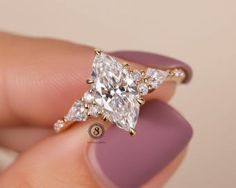 Pode incluir: Um anel de noivado dourado com um diamante de corte marquise como peça central, rodeado por diamantes menores. O anel tem uma banda delicada e é colocado sobre um fundo suave.