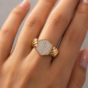 May include: A gold-colored ring with a hexagonal center filled with small, clear stones. The ring has a textured design on the sides, and is worn on a finger.
