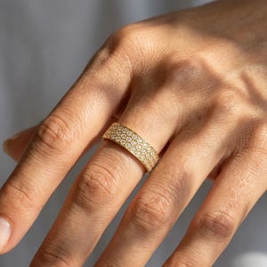 May include: A gold ring with a honeycomb design, set with small, clear gemstones. The ring is on a finger, and the light catches the facets of the stones, creating a sparkling effect. The ring is a band style, and the gold has a polished finish.