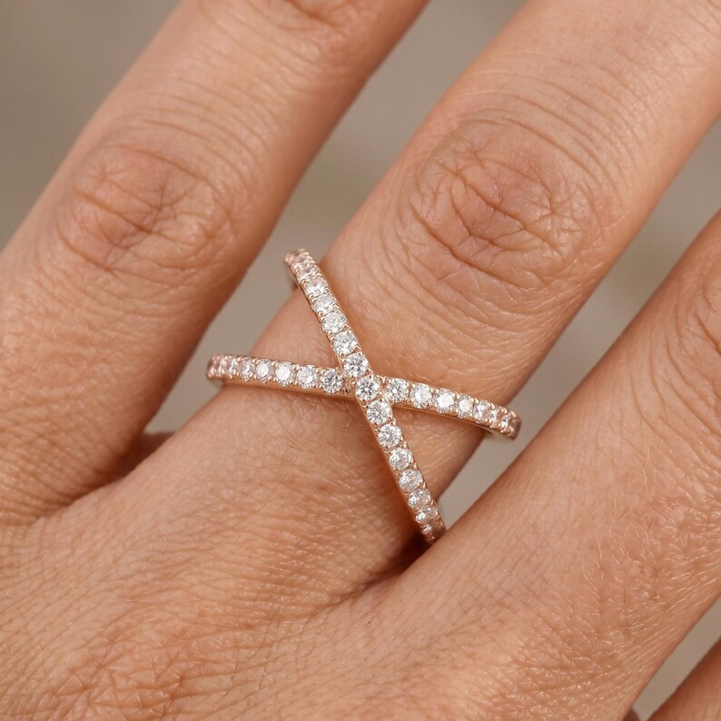 May include: Rose gold criss-cross ring with small, clear gemstones. The ring is set on a finger, showcasing the intricate design and sparkle of the stones. The band crosses over itself, creating an X shape.