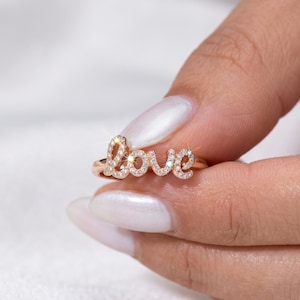 May include: A rose gold ring spelling out the word "love" in a cursive font. The letters are encrusted with small, sparkling clear gemstones. The ring is held up against a soft, white background.