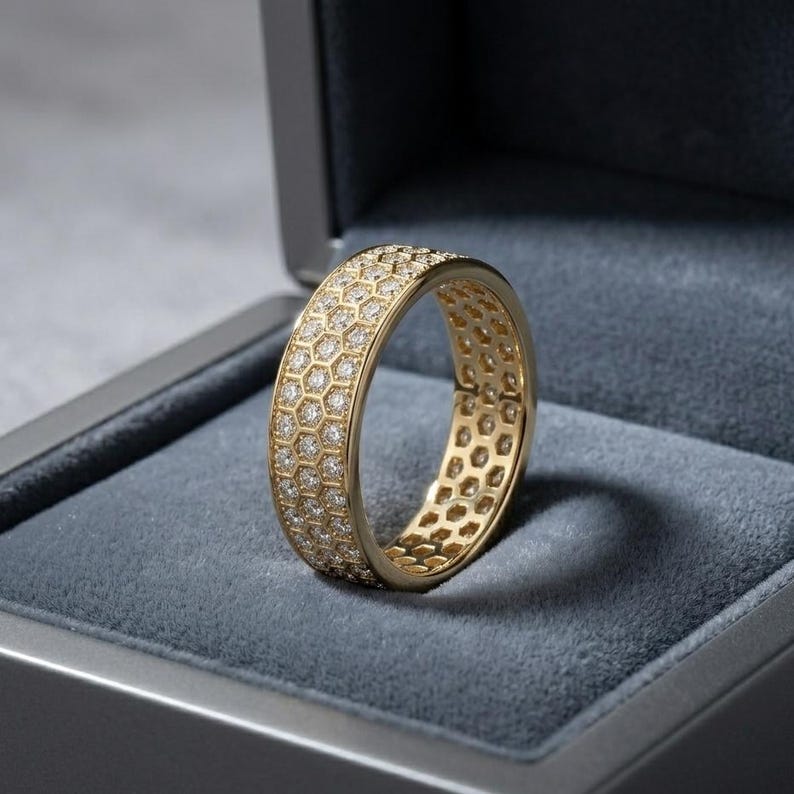 May include: A gold ring with a honeycomb design, encrusted with small, sparkling diamonds. The ring is displayed inside a gray jewelry box, showcasing its intricate details and luxurious appearance. The ring's band is approximately 0.5 inches wide.
