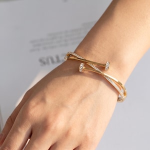 Puede incluir: Un brazalete dorado con un diseño inspirado en un clavo con dos extremos cruzados. La superficie del brazalete está adornada con pequeñas piedras claras y brillantes incrustadas a lo largo de la banda y en las cabezas planas del clavo.