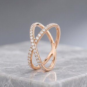 May include: Rose gold criss-cross ring with a double band design. The ring is encrusted with small, sparkling diamonds. The ring is set on a gray marble surface.