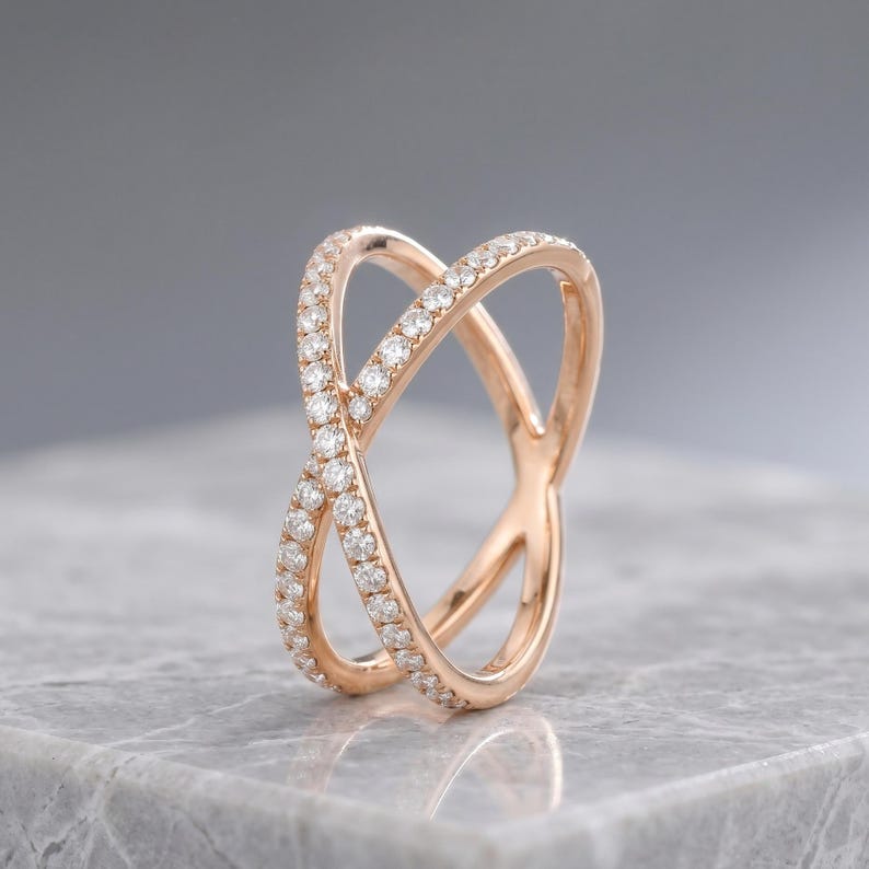 May include: Rose gold criss-cross ring with a double band design. The ring is encrusted with small, sparkling diamonds. The ring is set on a gray marble surface.