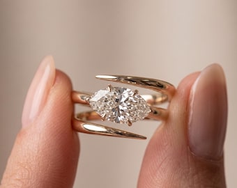 May include: A bypass ring crafted from polished rose gold, featuring a central marquise-cut white gemstone held by four prongs. The band consists of two sweeping, tapered arms that overlap slightly, creating an open, asymmetrical silhouette.