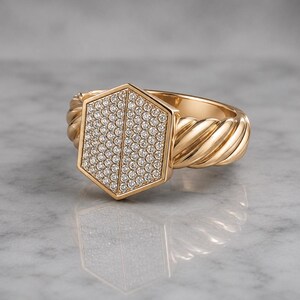 May include: A gold ring with a hexagonal face encrusted with small, sparkling stones. The ring's band features a twisted rope design. The ring is set against a light, reflective surface.