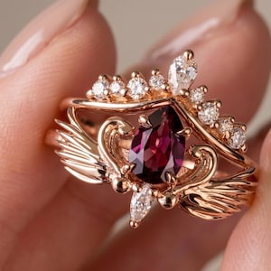 May include: A rose gold ring set featuring a central pear-shaped deep red gemstone flanked by stylized metal wings. The band is accented with a marquise-cut stone at the base and a curved crown-like topper set with multiple small round clear stones.