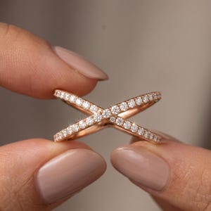 May include: Rose gold criss-cross ring with a pave of small, sparkling diamonds. The ring's design features two bands that intersect, creating an X shape. The ring is held between two fingers against a neutral background.