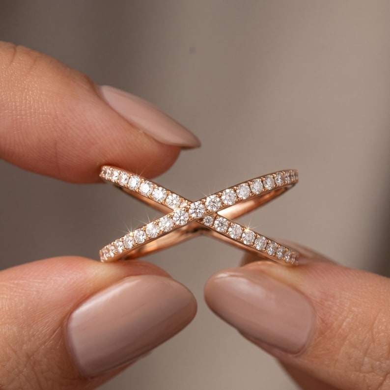May include: Rose gold criss-cross ring with a pave of small, sparkling diamonds. The ring's design features two bands that intersect, creating an X shape. The ring is held between two fingers against a neutral background.
