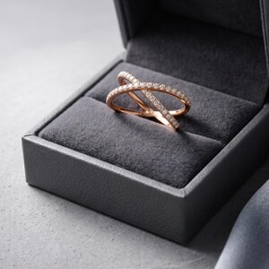 May include: Rose gold criss-cross ring with small, clear gemstones set in a gray jewelry box. The ring's design features two bands that intersect, creating an X shape. The box is open, revealing the ring's intricate details.
