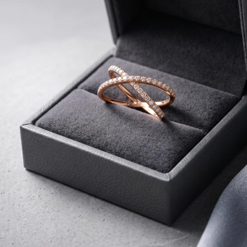 May include: Rose gold criss-cross ring with small, clear gemstones set in a gray jewelry box. The ring's design features two bands that intersect, creating an X shape. The box is open, revealing the ring's intricate details.