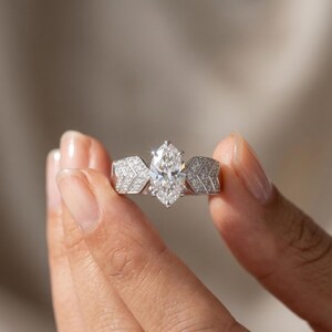 May include: A marquise-cut diamond engagement ring. The ring features a large, central diamond and smaller diamonds set in a chevron design on the band. The ring is made of white metal, likely platinum or white gold.