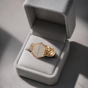 May include: A gold ring with a hexagonal face encrusted with small, sparkling stones. The ring's band features a twisted rope design. The ring is presented in an open, gray velvet jewelry box, set against a neutral background.