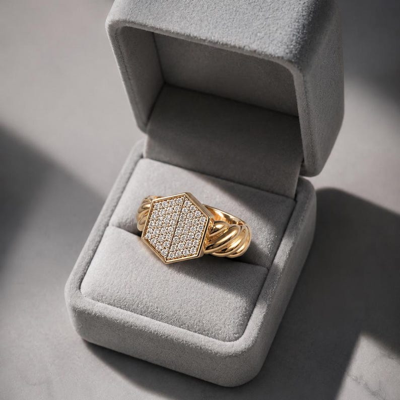 May include: A gold ring with a hexagonal face encrusted with small, sparkling stones. The ring's band features a twisted rope design. The ring is presented in an open, gray velvet jewelry box, set against a neutral background.