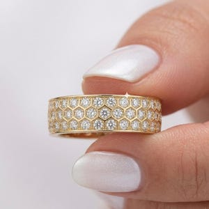 May include: A gold ring with a honeycomb design, set with numerous small, sparkling diamonds. The ring is held up against a white background, showcasing its intricate details and brilliant shine. The band is wide, and the diamonds are evenly spaced.