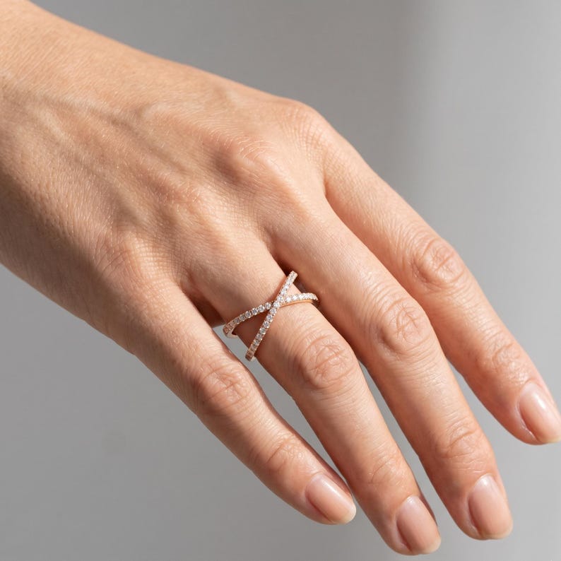 May include: A rose gold criss-cross ring with small, clear gemstones. The ring is on a hand with a neutral background. The ring's design features two bands that intersect, creating an X shape. The gemstones are evenly spaced along the bands.