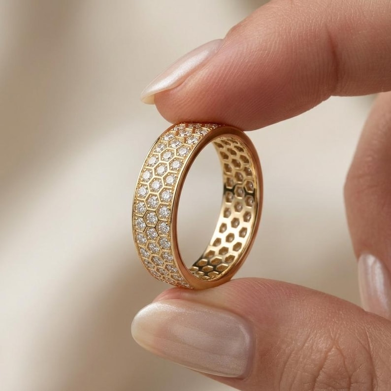 May include: A gold ring with a honeycomb design, encrusted with small, sparkling gemstones. The ring is held between two fingers, showcasing its intricate details and radiant appearance. The ring is a piece of jewelry.