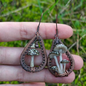 以下が含まれることがあります： 手作りの銅線イヤリング。各イヤリングは、ミニチュアキノコのデザインのしずく型のフレームが特徴です。キノコは帽子に白い斑点があり、緑色の背景に設定されています。