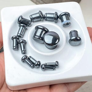 Op de afbeelding: Een verzameling kleine, gladde, donkergrijze metalen oorpluggen met een gepolijste afwerking. De set bevat verschillende maten cilindrische pluggen met afgeronde, platte uiteinden, gerangschikt op een wit vierkant presenteerblad met een inkeping.