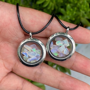 May include: Two circular silver-toned metal lockets suspended on black cords. Each locket features a clear glass front and back, containing multiple small, iridescent, white-toned opal-like stones that display flashes of blue, green, and pink.