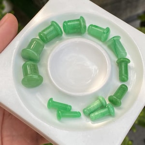 Puede incluir: Una colección de diez dilatadores de oreja cilíndricos de color verde claro con extremos acampanados, dispuestos en un patrón circular sobre una bandeja cuadrada blanca. Cada pieza presenta una superficie lisa y semitranslúcida.