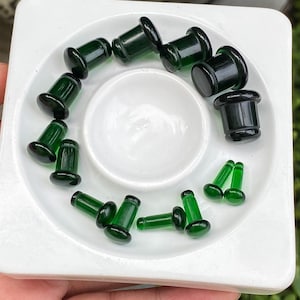 Puede incluir: Una colección de dilatadores de oreja de vidrio verde oscuro y pendientes más pequeños de vidrio verde dispuestos en un patrón circular sobre una bandeja cuadrada blanca. Los dilatadores presentan un diseño acampanado y pulido.