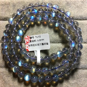 May include: A triple-strand bracelet made of translucent, round labradorite beads with iridescent blue flashes. A white tag attached to the beads displays the text: TU10, 6.5MM, and Chinese characters indicating a natural labradorite triple-strand bracelet.