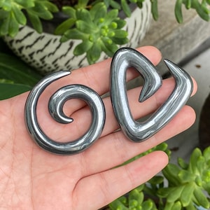 Può includere: Due dilatatori per orecchie color grigio scuro con finitura metallica liscia e lucida. Uno presenta un design a spirale circolare, mentre l'altro ha una forma triangolare simile a un cuore con una punta curva, entrambi realizzati in materiale solido.